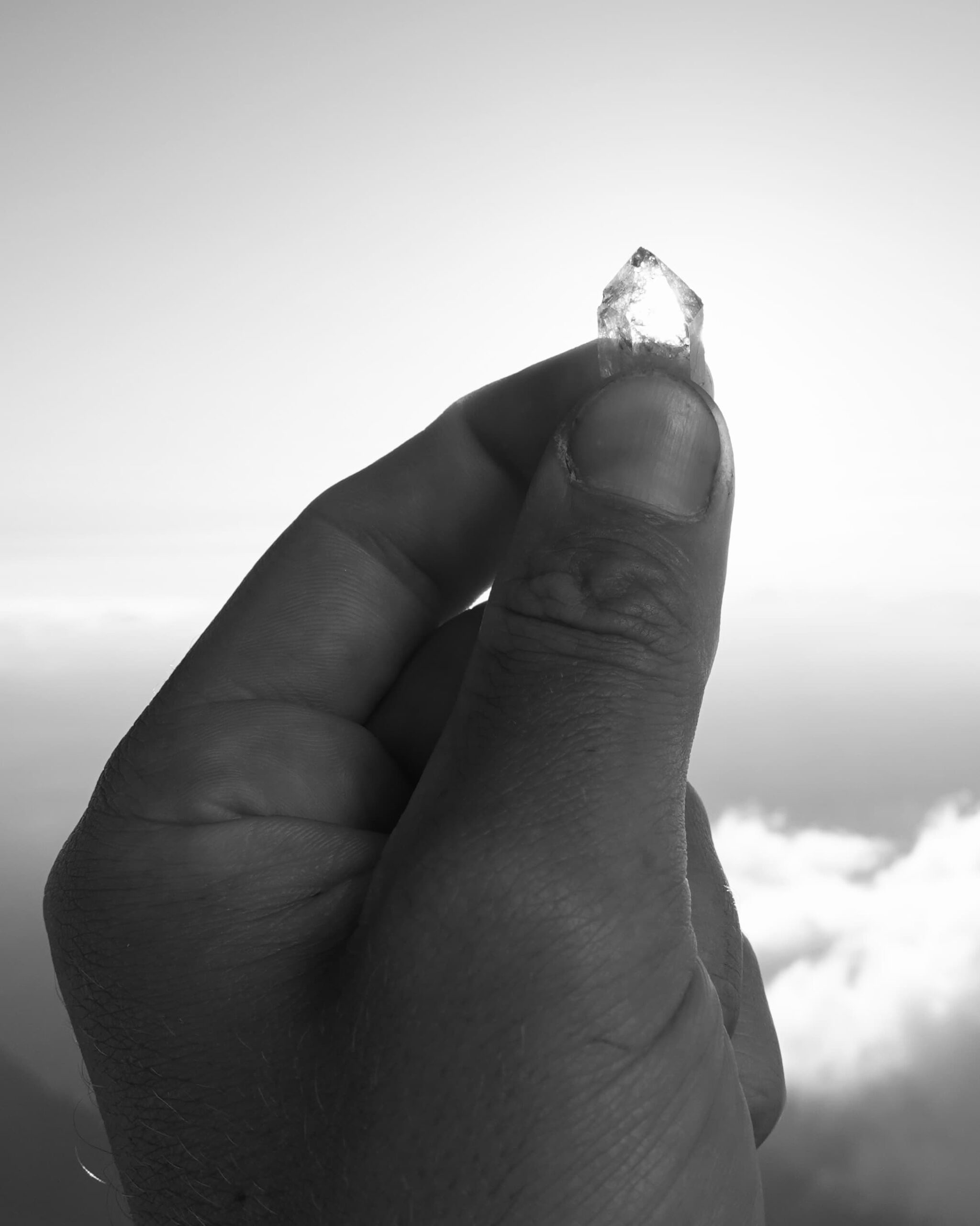 Eine Nahaufnahme einer Hand, die einen kleinen, durchscheinenden Kristall vor einem strahlend blauen Himmel hält, im Hintergrund sind Wolken zu sehen. Das Bild ist in Schwarz-Weiß gehalten.