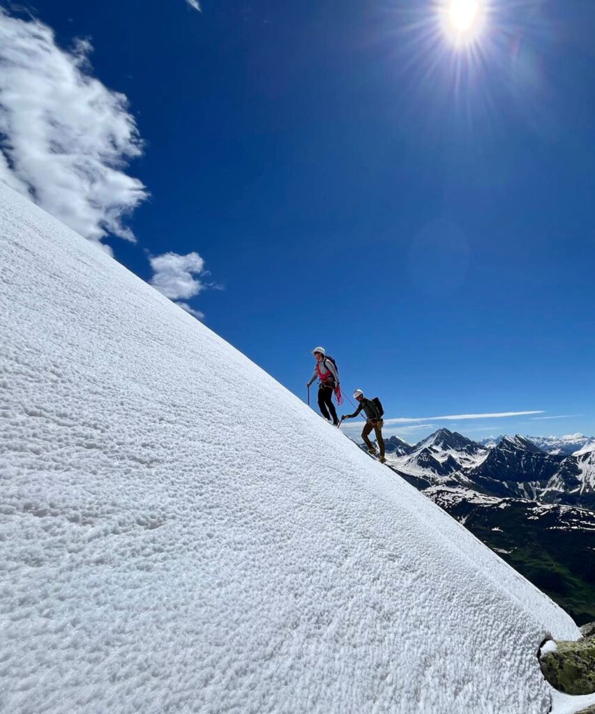 Zwei Personen begeben sich auf geführte Bergtouren und steigen unter strahlendem Sonnenschein und bei strahlend blauem Himmel einen steilen, verschneiten Hang hinauf. Im Hintergrund sind schneebedeckte Berge und ein grünes Tal zu sehen.