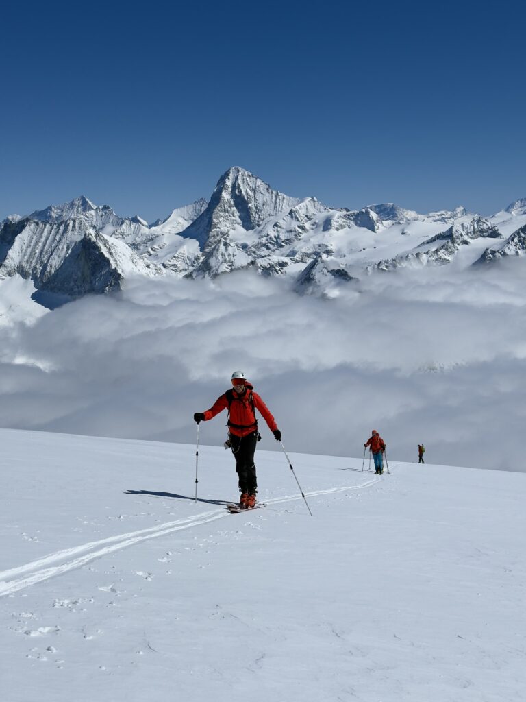 Drei Personen in Winterkleidung fahren auf einem verschneiten Berghang bergauf und nehmen an geführten Bergtouren teil, im Hintergrund sind schneebedeckte Gipfel und Wolken vor einem strahlend blauen Himmel zu sehen.