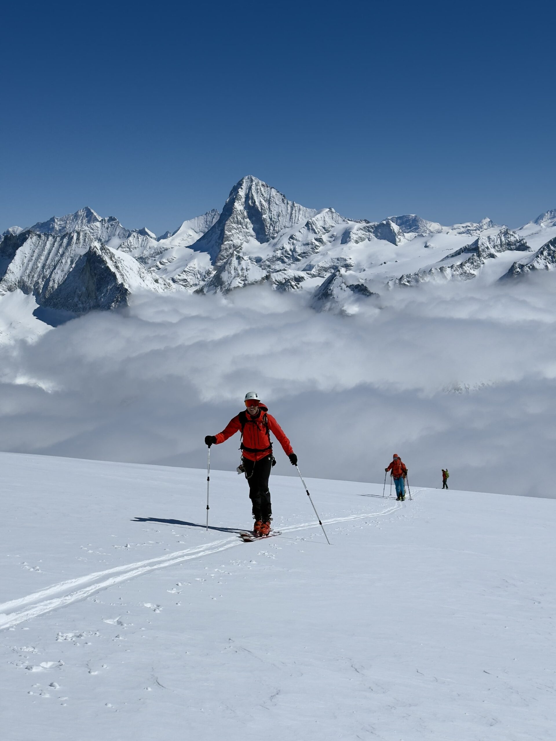 Drei Personen in Winterkleidung fahren auf einem verschneiten Berghang bergauf und nehmen an geführten Bergtouren teil, im Hintergrund sind schneebedeckte Gipfel und Wolken vor einem strahlend blauen Himmel zu sehen.