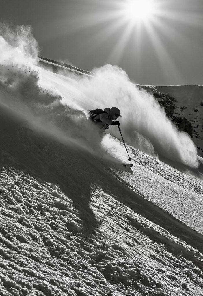 Ein Skifahrer saust auf frischem Pulverschnee den Hang hinunter, wirbelt dabei im strahlenden Sonnenlicht Schnee auf – mit dramatischen Schatten und einer Bergkulisse im Hintergrund: eine aufregende Szene, die an Erlebnisse bei geführten Bergtouren erinnert.