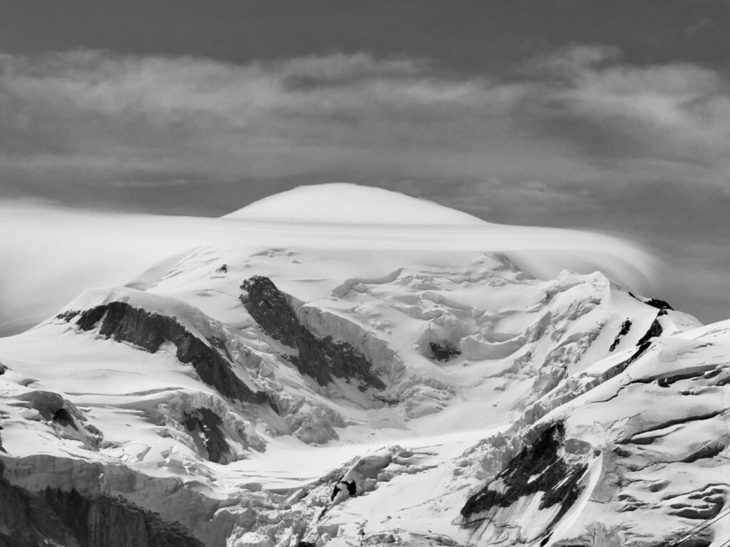 Ein schneebedeckter Berggipfel, um den sich unter einem teilweise bewölkten Himmel dramatische Wolken wabern, eingefangen in Schwarz-Weiß – eine atemberaubende Szene, die an die Landschaften erinnert, die man auf geführten Bergtouren in den Alpen erkundet.