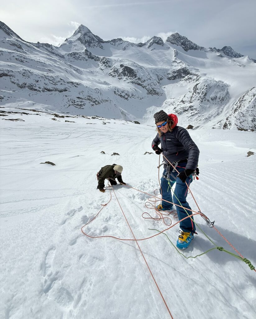 Zwei Bergsteiger in Kletterausrüstung befinden sich auf einem verschneiten Hang und genießen das Abenteuer einer geführten Bergtour. Eine Person steht lächelnd im Vordergrund, während eine andere hinter ihr im Schnee hockt. Vor dem Hintergrund weitläufiger, verschneiter Berggipfel sind sie durch Kletterseile miteinander verbunden.
