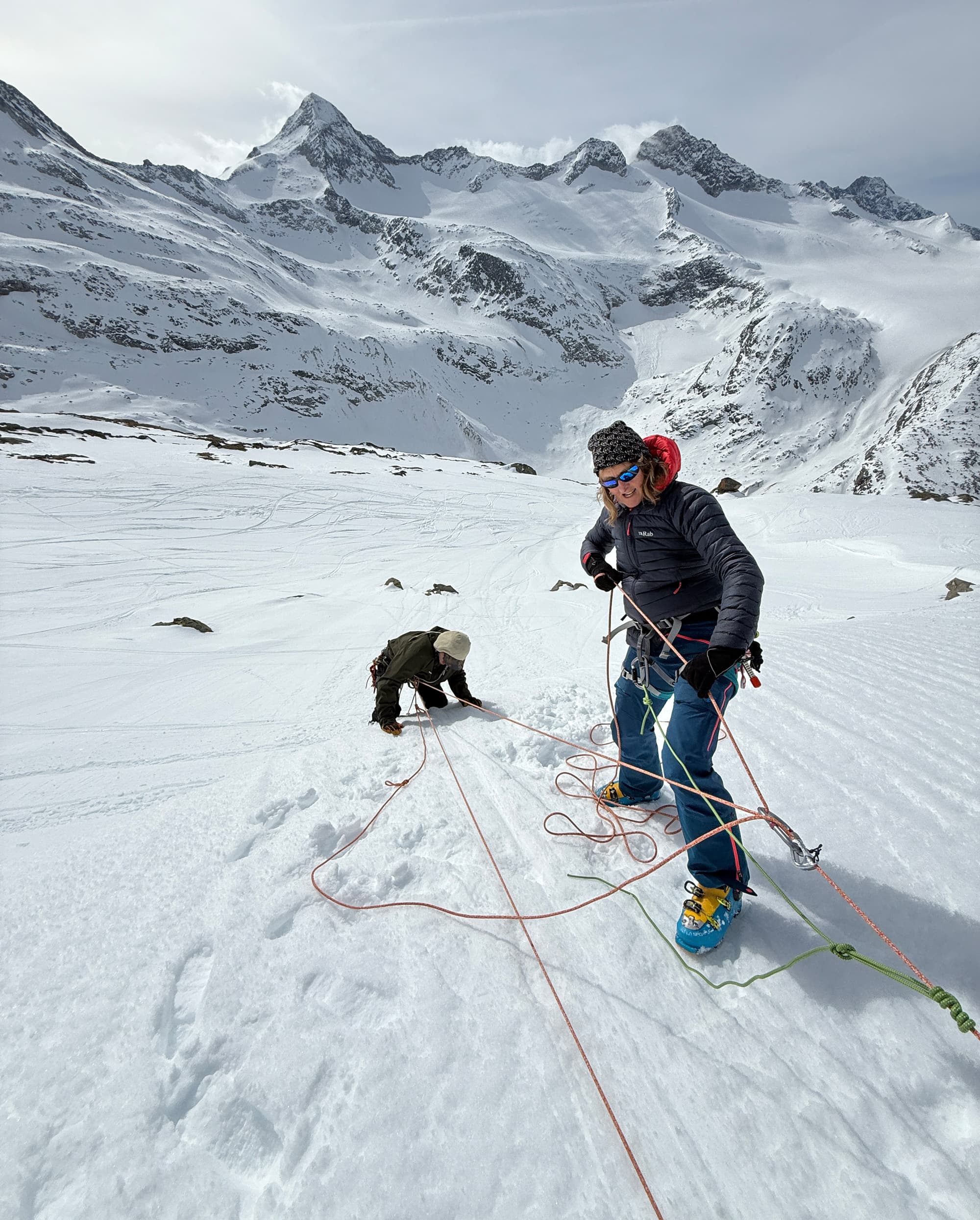 Zwei Bergsteiger in Kletterausrüstung befinden sich auf einem verschneiten Hang und genießen das Abenteuer einer geführten Bergtour. Eine Person steht lächelnd im Vordergrund, während eine andere hinter ihr im Schnee hockt. Vor dem Hintergrund weitläufiger, verschneiter Berggipfel sind sie durch Kletterseile miteinander verbunden.