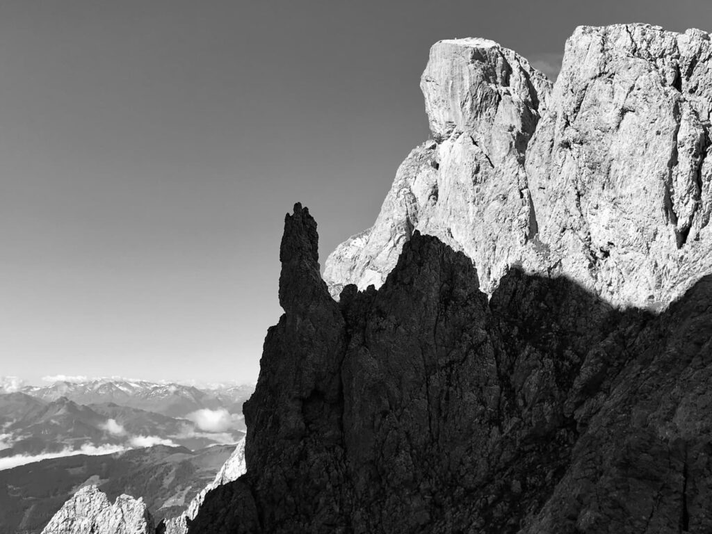Zerklüftete Felswände im Wechsel von grellem Sonnenlicht und Schatten, mit fernen Bergen und Wolken, die am Horizont unter einem klaren Himmel zu sehen sind – ein stimmungsvolles Schwarz-Weiß-Foto, das an Landschaften erinnert, die man auf geführten Bergtouren erkundet hat.