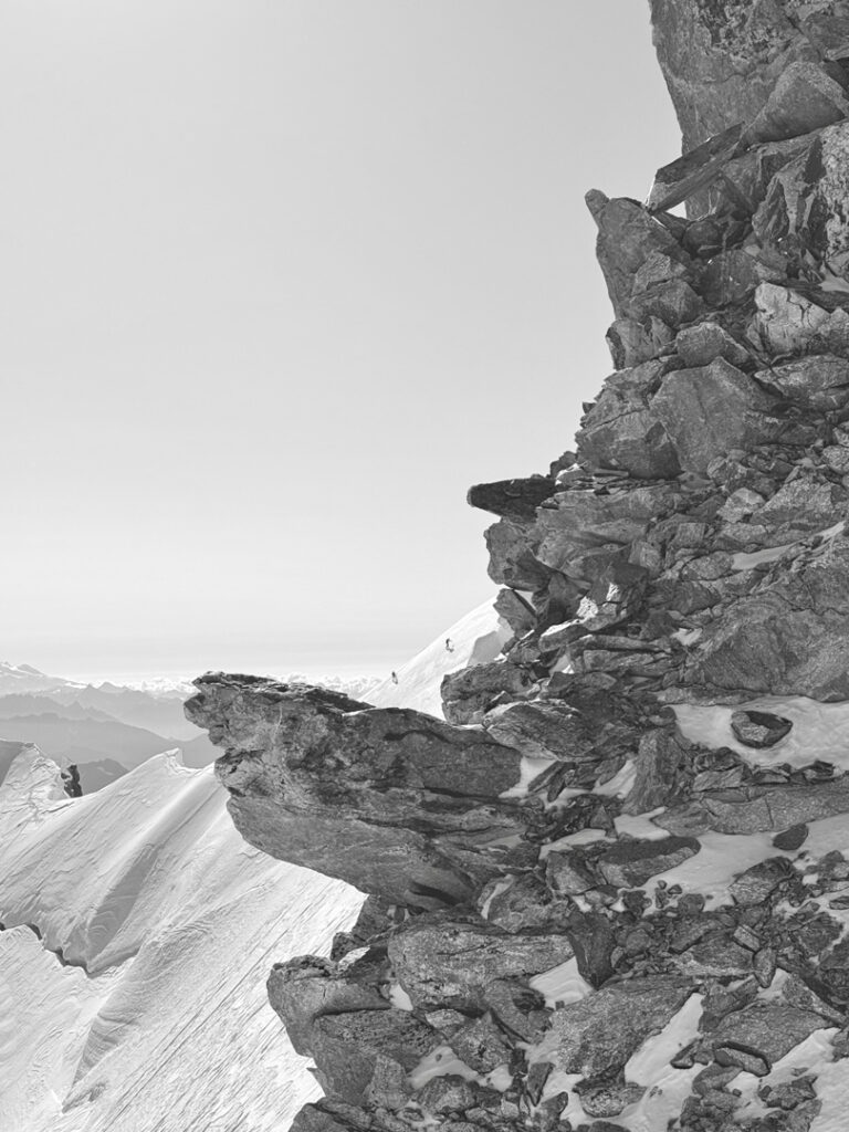 Von der rechten Seite erstreckt sich eine schneebedeckte, felsige Klippe mit zerklüfteten Kanten; im Hintergrund sind verschneite Berge in der Ferne und ein klarer Himmel zu sehen, was eine atemberaubende Kulisse bildet, die sich ideal für geführte Bergtouren eignet.