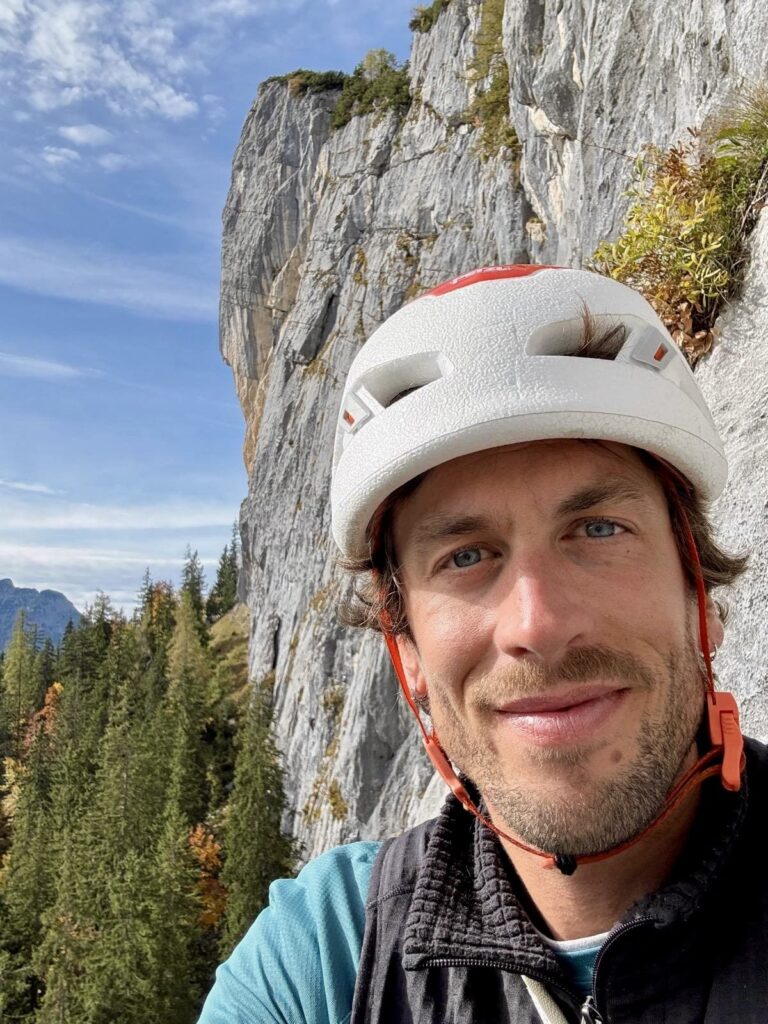 Ein Mann mit einem weißen Kletterhelm lächelt für ein Selfie vor dem Hintergrund einer felsigen Klippe und Kiefern unter einem teilweise bewölkten Himmel und hält damit einen abenteuerlichen Moment fest, der jedem ARE YOU SUMMIT-Bergführer alle Ehre machen würde.